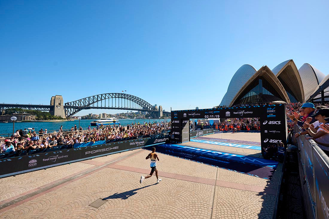 Arrivée spectaculaire du TCS Sydney Marathon avec l'Opéra et le Harbour Bridge en arrière-plan, un coureur franchissant la ligne sous les applaudissements de la foule.